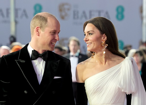 Princesa Kate Middleton usa cabelo partido ao meio e brinco maxi de flores douradas da Zar. Ela usa um longo vestido branco e luvas pretas. Ao lado está o príncipe William com um smoking preto e gravata borboleta. Ele é um homem branco e careca- Metrópoles 