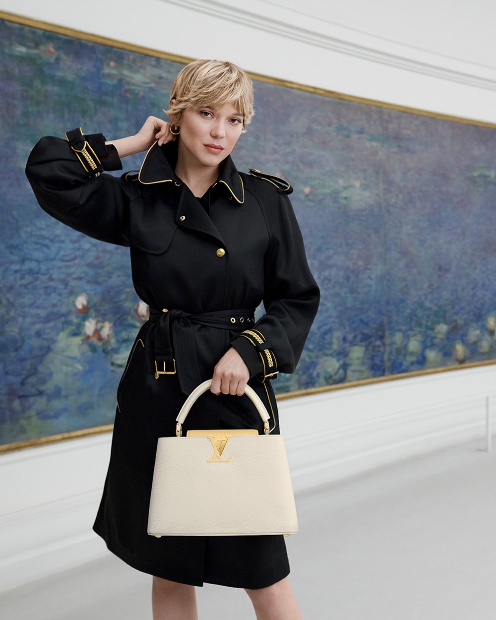 A atriz Léa Seydoux, uma mulher branca e jovem, de cabelo loiro liso e curto, posa em campanha da marca Louis Vuitton. Ela está sentada em um banco, dentro de um museu, e usa um top preto, uma saia branca, um casaco preto e a bolsa Capucines, de couro, da marca. - Metrópoles