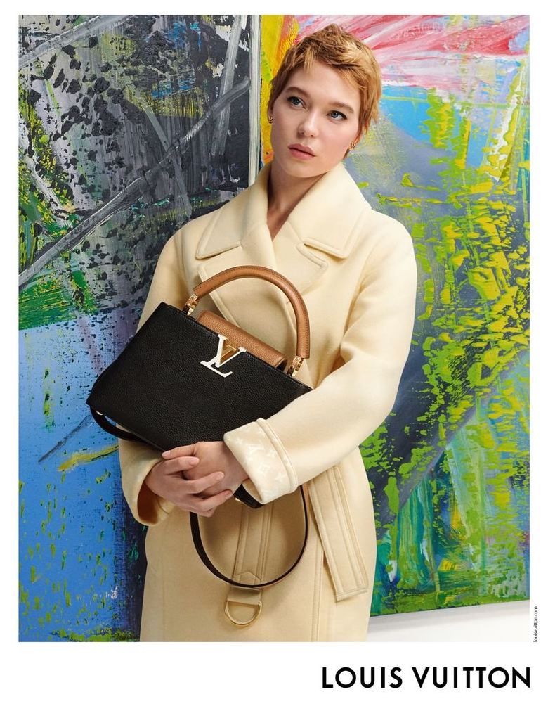 A atriz Léa Seydoux, uma mulher branca e jovem, de cabelo loiro liso e curto, posa em campanha da marca Louis Vuitton. Ela está encostada em uma obra de arte, dentro de um museu, e usa um casaco bege e a bolsa Capucines, de couro, da marca. - Metrópoles