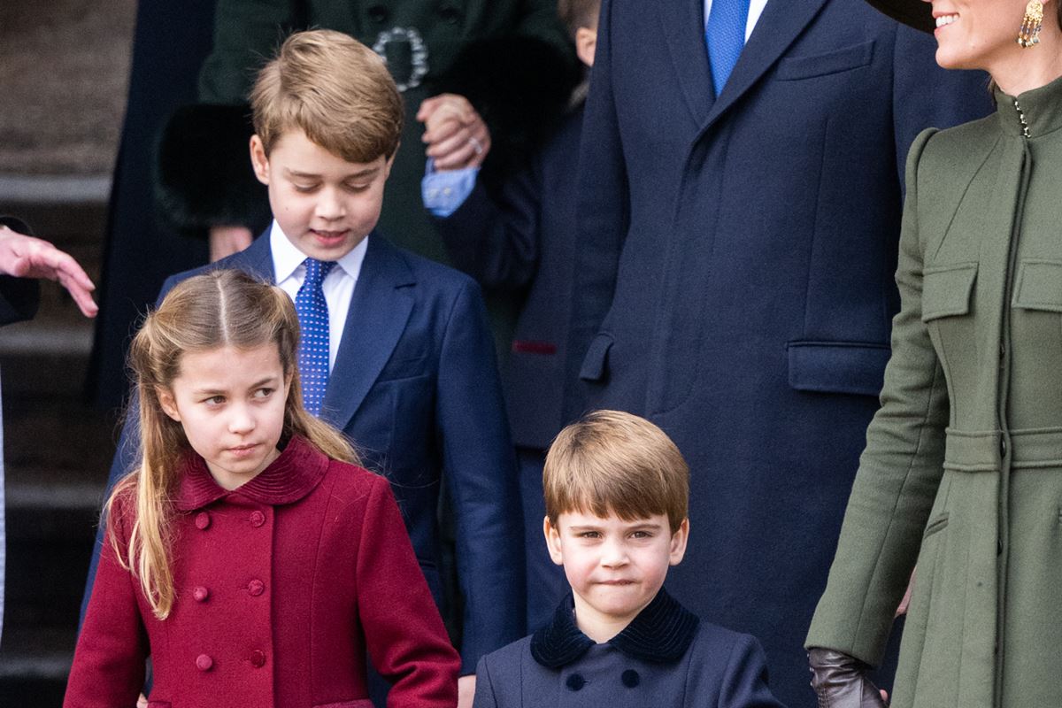 Foto colorida de dois meninos e uma menina, com casaco vermelho. Os dois meninos estão com roupas pretas. Eles descem uma escada ao lado de uma mulher com roupa verde militar - Metrópoles