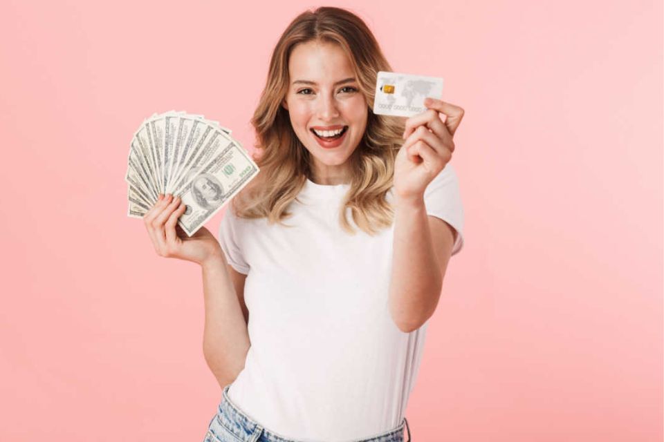 Mulher loira segurando dinheiro e cartão com uma blusa branca e fundo rosa