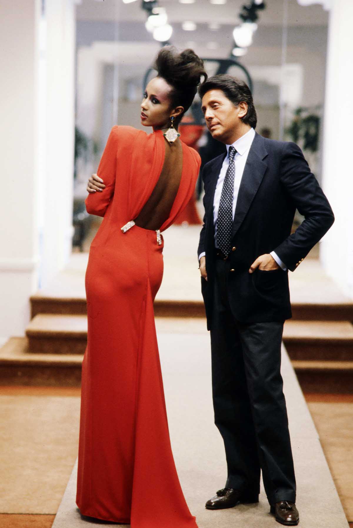 O estilista Valentino Garavani com a modelo Iman Abdulmajid em 1984. Ela é uma mulher negra e jovem e usa um vestido longo vermelho. Ele é um homem branco, de meia idade, que usa terno e gravata. - Metrópoles