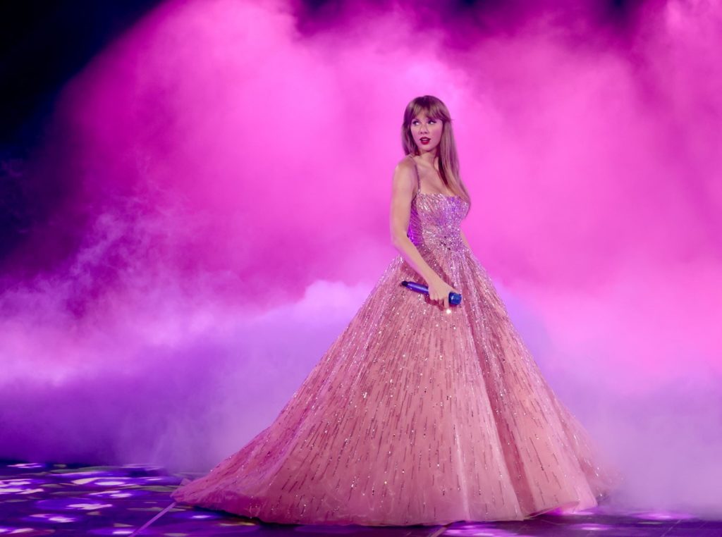 A cantora Taylor Swift, uma mulher jovem e branca, de cabelo loiro liso. Ela está no palco, na turnê The Eras Tour, e usa um branco de tule, com o bustiê rosa bordado, da marca Zuhair Murad. - Metrópoles