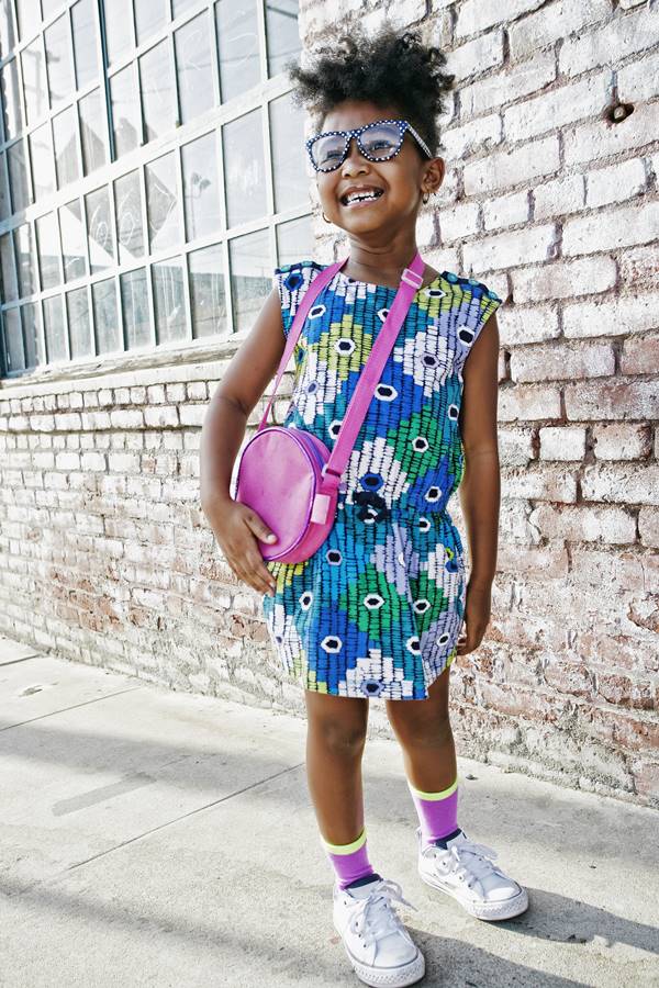 Criança na rua posando para foto com roupas coloridas. Ela está usando óculos de sol e apoiando uma das mãos em uma bolsa - Metrópoles