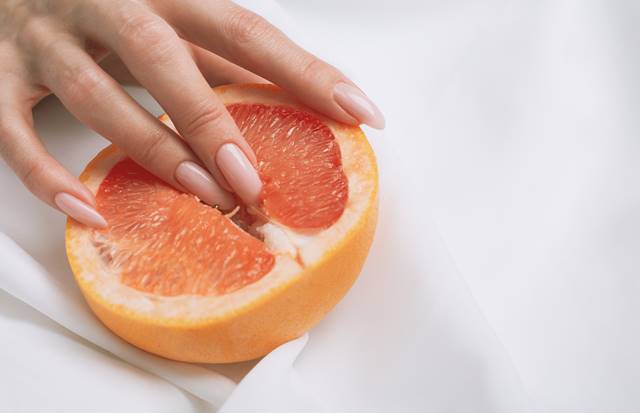 Dedos femininos com unhas grandes dentro de uma toranja em um fundo branco - Metrópoles