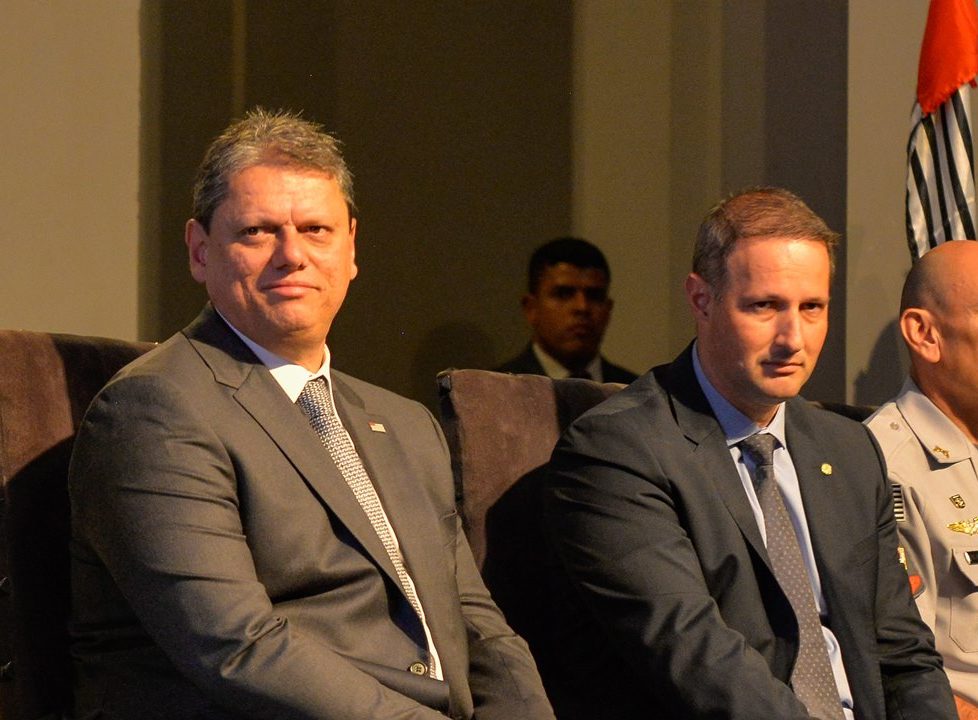 Imagem colorida do governador de São Paulo Tarcísio de Freitas sentado ao lado do secretário de Segurança Pública Guilherme Derrite. Os dois estão vestidos de terno e gravata