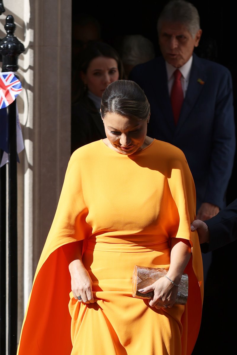 Foto colorida de mulher com um vestido laranja e uma bolsa nas mãos. Ela sai de um portão - Metrópoles