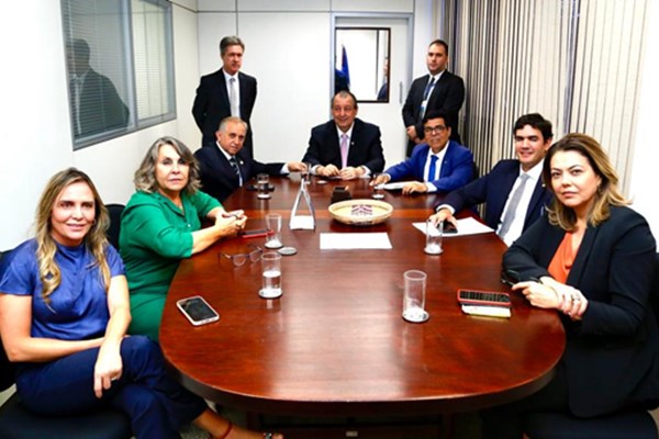 Fotografia colorida mostra grupo de sete pessoas sentados à mesa e dois homens em pé, ao fundo