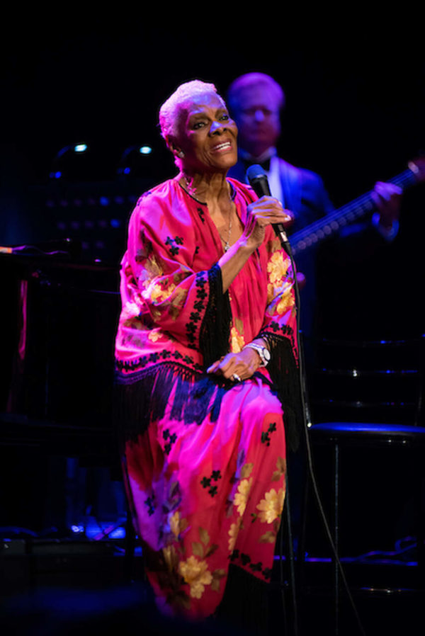 Na imagem com cor, uma mulher negra cantando ao lado de um piano usando vestido vermelho - Metrópoles