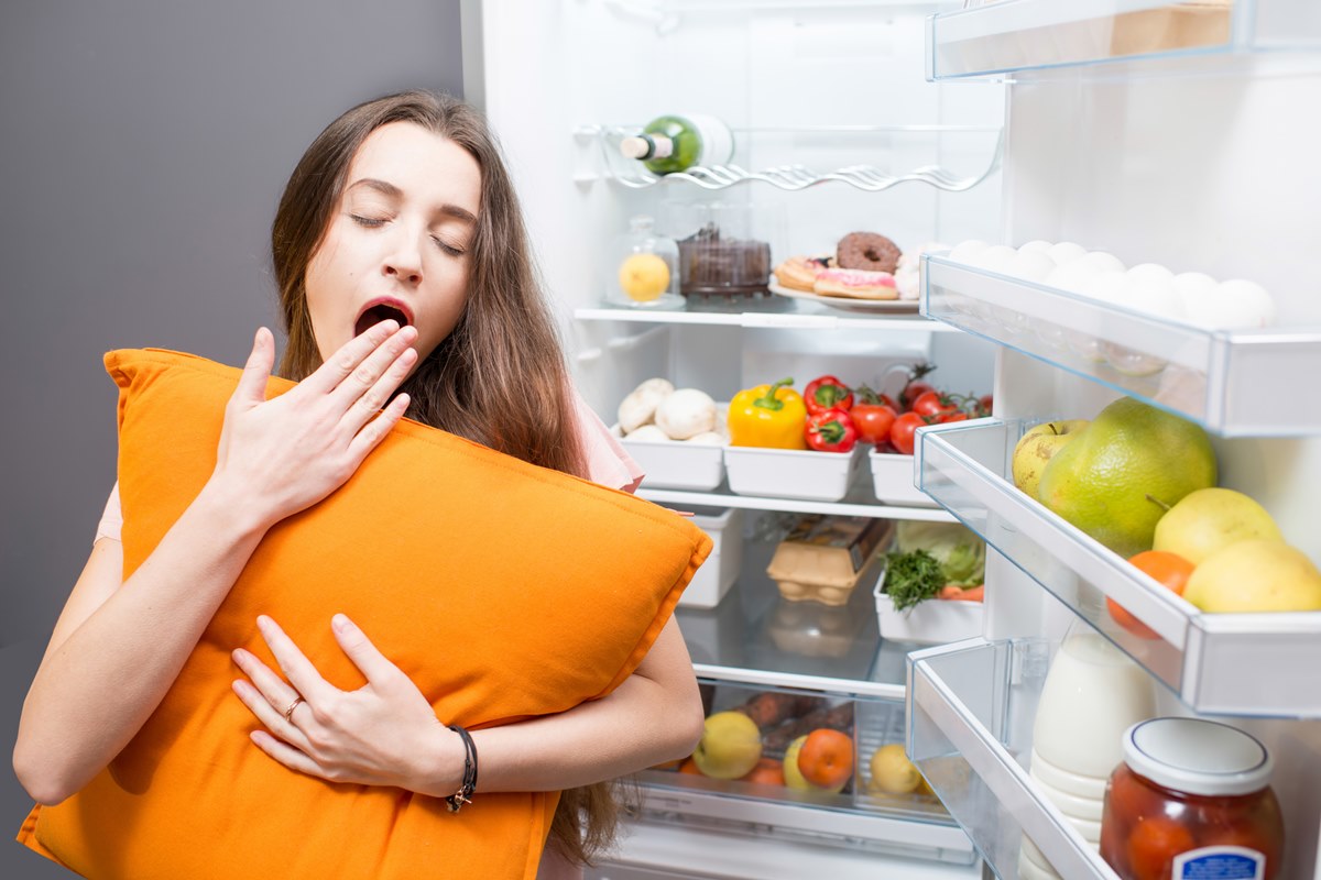 Foto colorida de mulher com almofada em frente a uma geladeira cheia de alimentos - Metrópoles