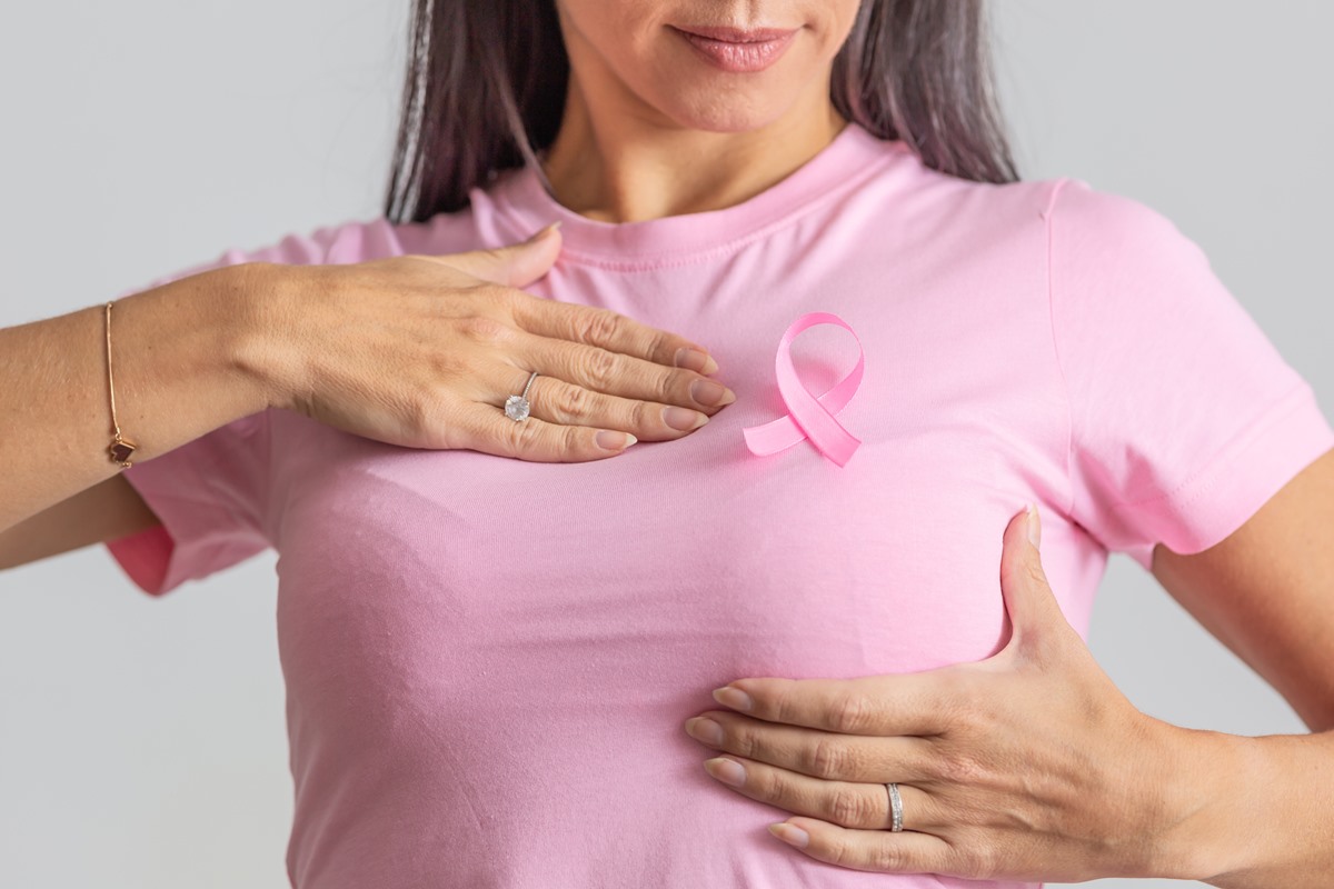 Foto colorida de mulher de blusa rosa apalpando os seios em campanha contra o câncer de mama - Metrópoles