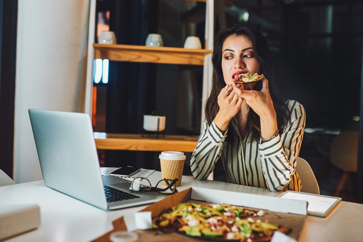 Foto colorida de mulher branca comendo pizza em frente ao notebook - Metrópoles