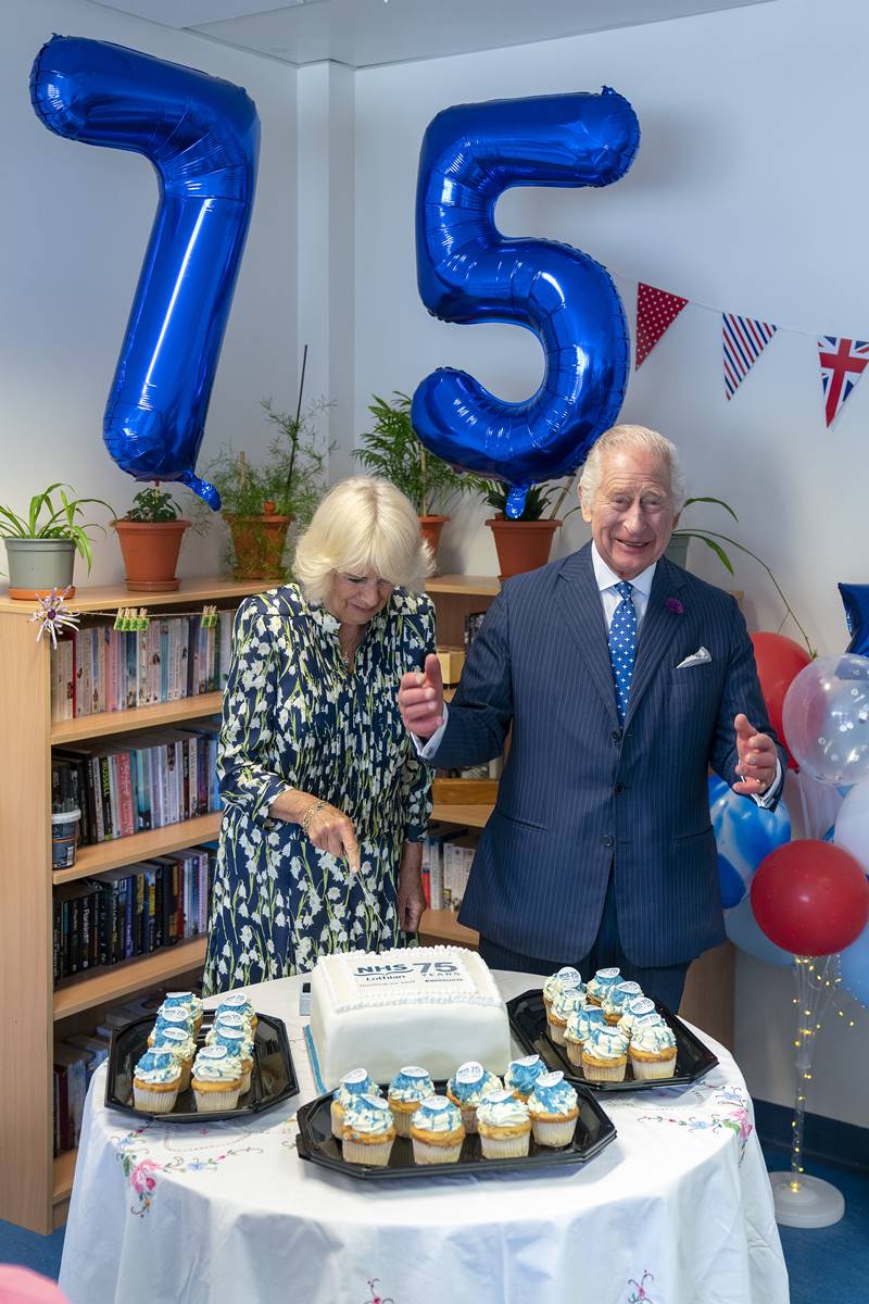 Foto colorida de mulher idosa e branca cortando um bolo ao lado de um homem idoso e branco sorridente. Atrás, há dois balões com azuis presos na parede com os números 7 e 5 - Metrópoles