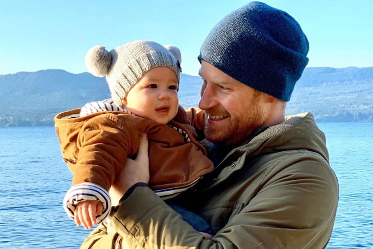 Foto colorida de bebê branco, com touca e casaco bege. Ele está em colo de homem branco, ruivo e com casaco verde-militar e touca azul. Ao fundo, há um rio - Metrópoles