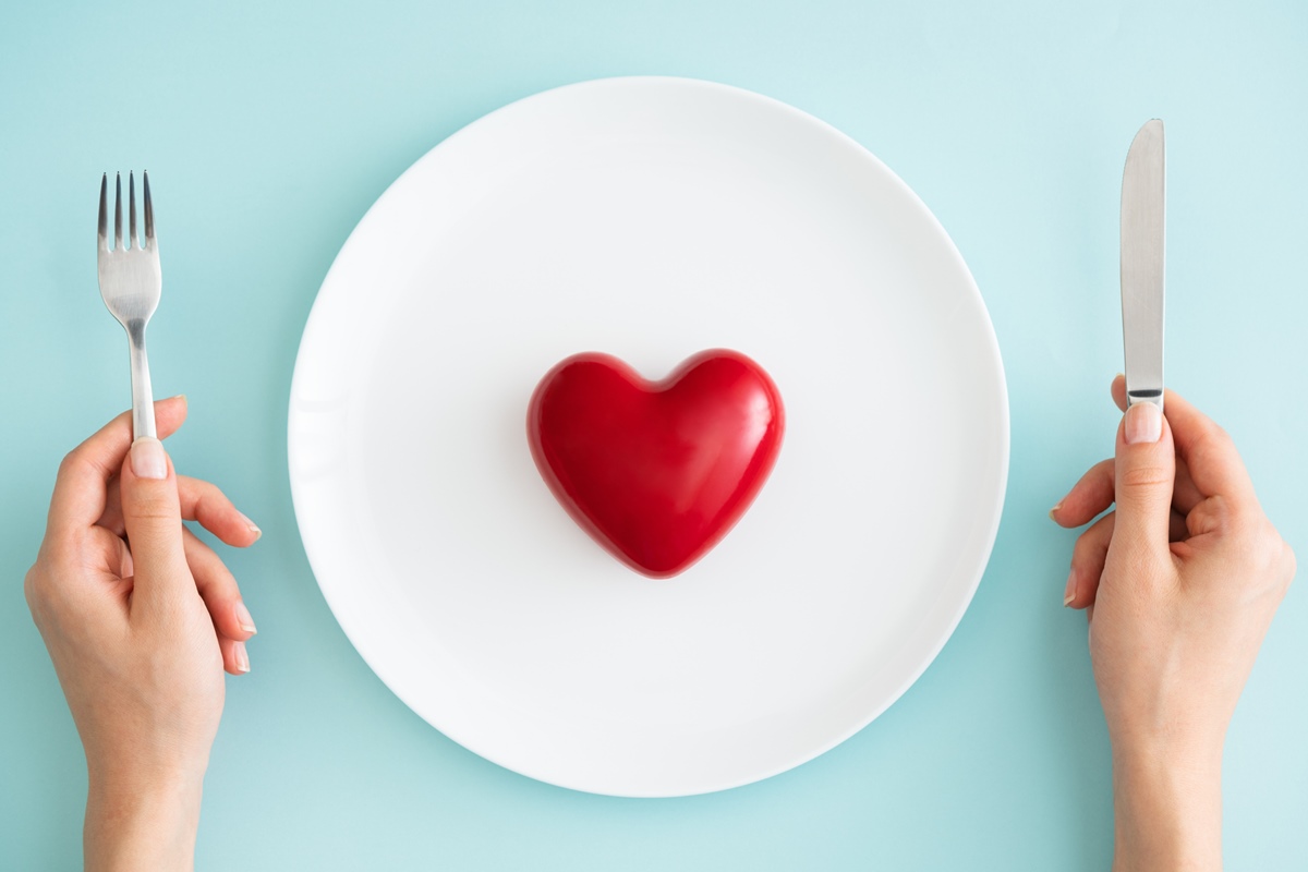 Foto colorida de um coração vermelho, dentro de um prato prato, ao lado está um par de mãos que segura um garfo e uma faca - Metrópoles