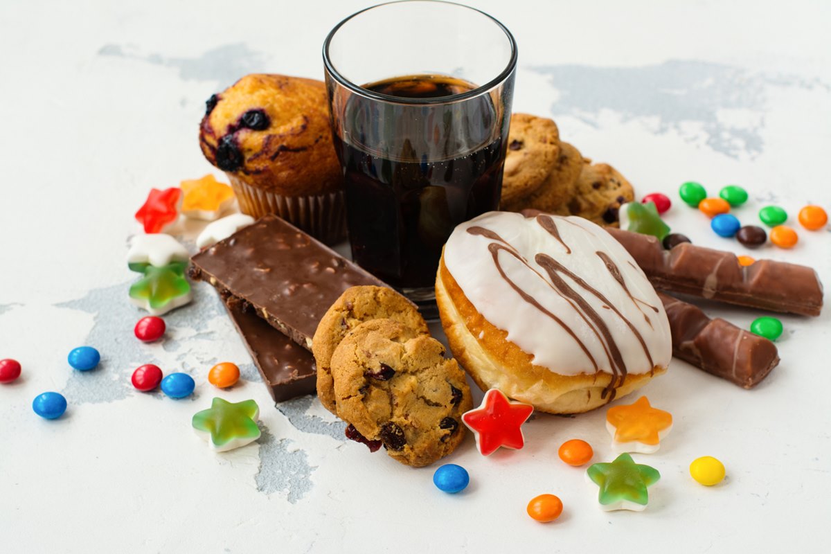 Foto colorida de alimentos, incluindo balinhas, barras de chocolate, copo de refrigerante, donuts, cookies e muffins - Metrópoles