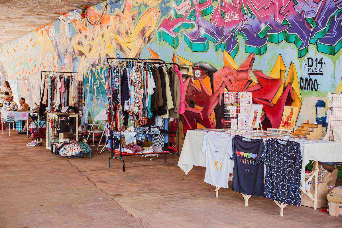 Na imagem com cor, araras com roupas e acessórios em frente a mural colorido - Metrópoles