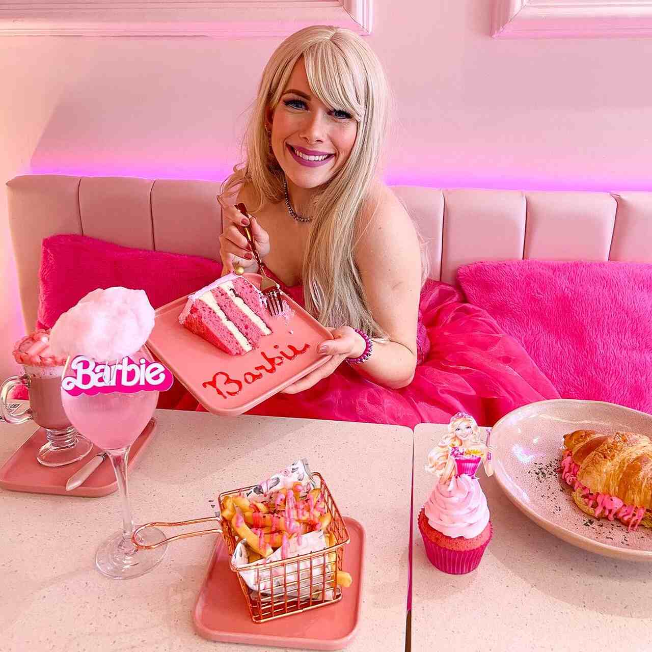 Na foto, uma mulher loi8ra sentada em uma mesa com comidas, segurando uma torta rosa - Metrópoles