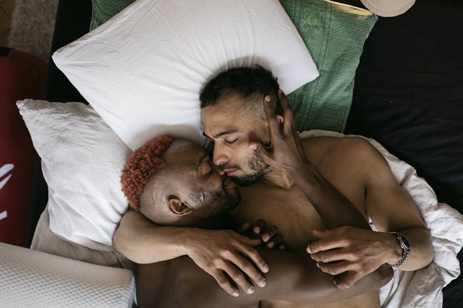 Foto colorida de um casal de dois homens deitados e abraçados na cama, com dois travesseiros e um lençol verde - Metrópoles