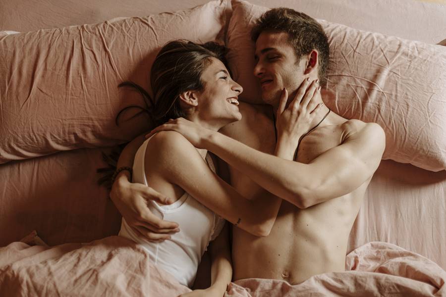 Foto colorida de cima de um casal com um homem e uma mulher deitados em uma cama de lençol rosa, sorrindo olhando um para o outro, abraçados - Metrópoles