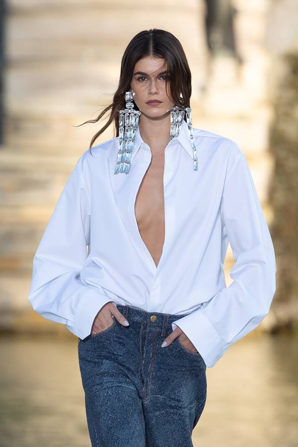Na passarela de moda, a modelo Kaia Gerber usa brinco grande com camisa branca com decote e calça jeans - Metrópoles