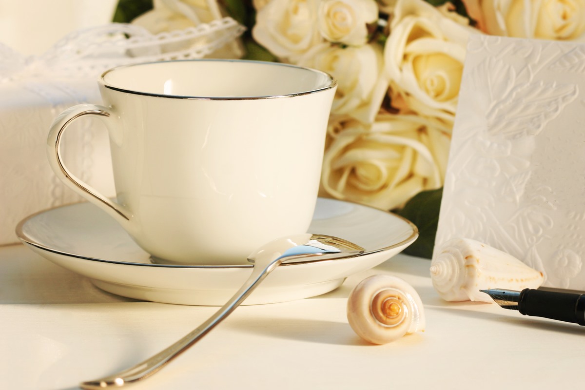 Foto colorida de xícara e pires com colher dourada em cima de uma mesa. Ao fundo, tem um buquê de rosas brancas. Também tem um papel perto das flores - Metrópoles