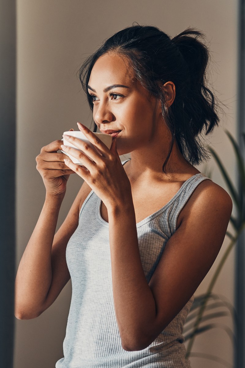 Foto colorida de mulher negra tomando algo dentro de uma xícara. Ela está com uma blusa regata cinza - Metrópoles