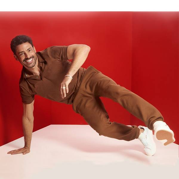 Cauã Reymond em campanha de moda. Em movimento, com apenas uma mão apoiada no chão, ele usa look marrom com calça e camisa com gola polo - Metrópoles