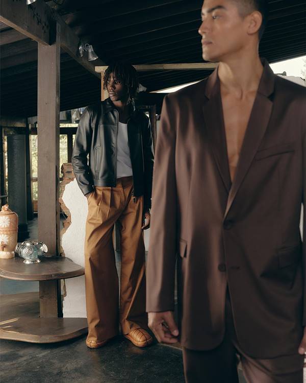 Campanha de moda com dois homens. Um deles, à frente, usa blazer marrom. O outro, que está atrás, está com camiseta branca, jaqueta preta, além de calça e calçado marrons - Metrópoles