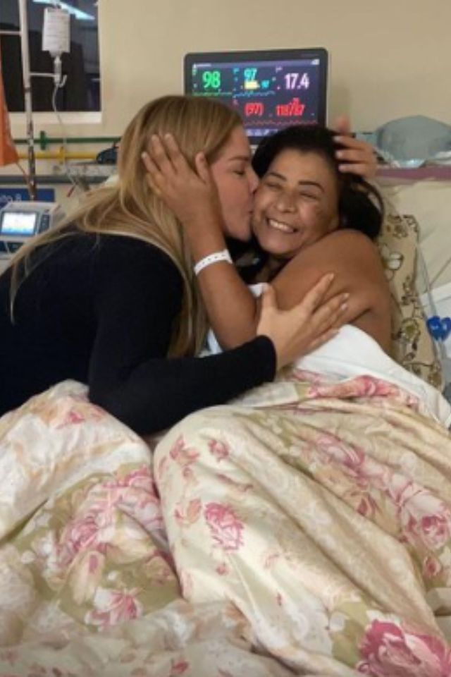 Foto colorida de duas mulheres, uma em cima de uma cama hospitalar e outra beijando o seu rosto - Metrópoles