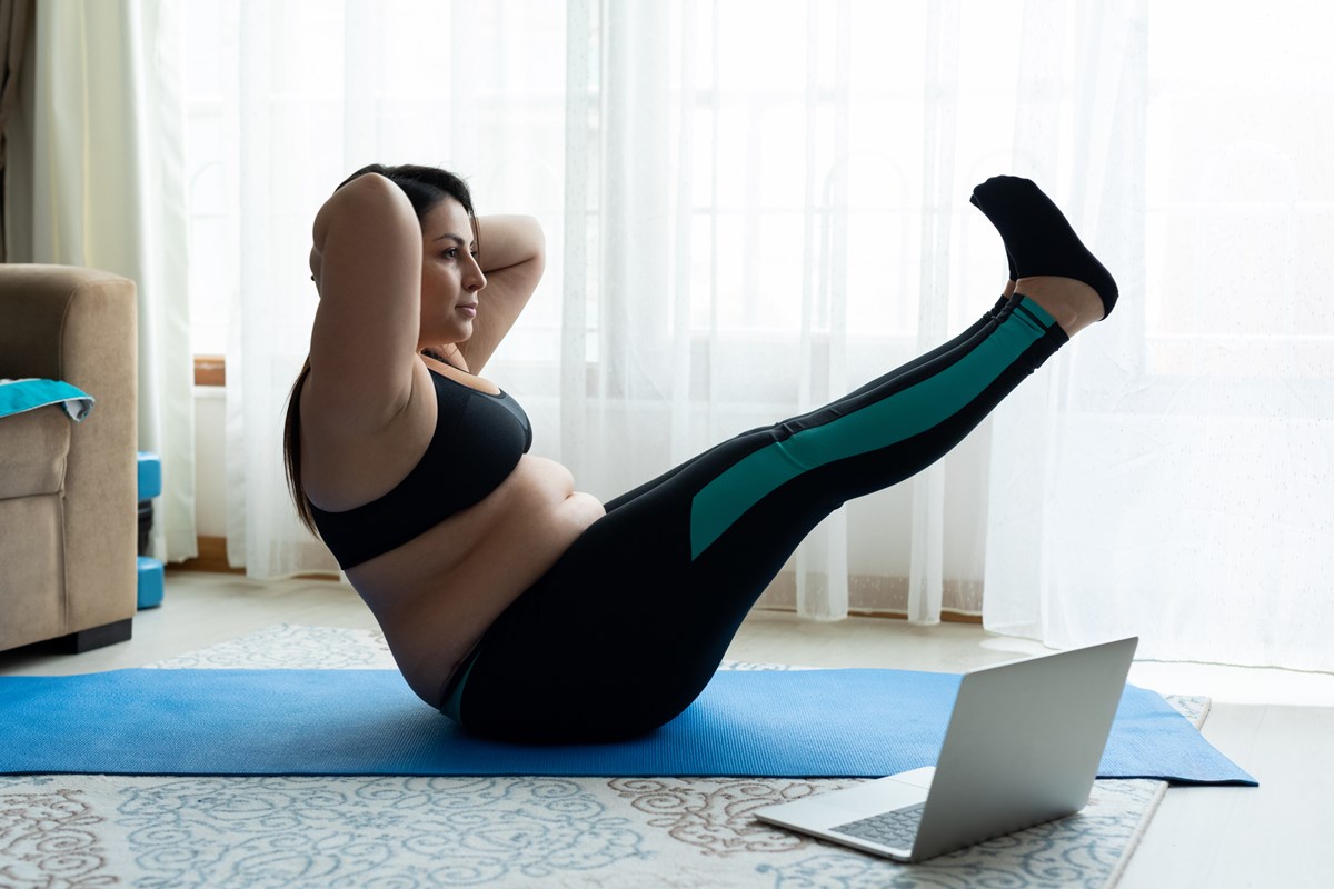 Foto colorida de mulher branca fazendo exercício com o próprio corpo. Ela está em um tapete de ioga e olha para um computador - Metrópoles