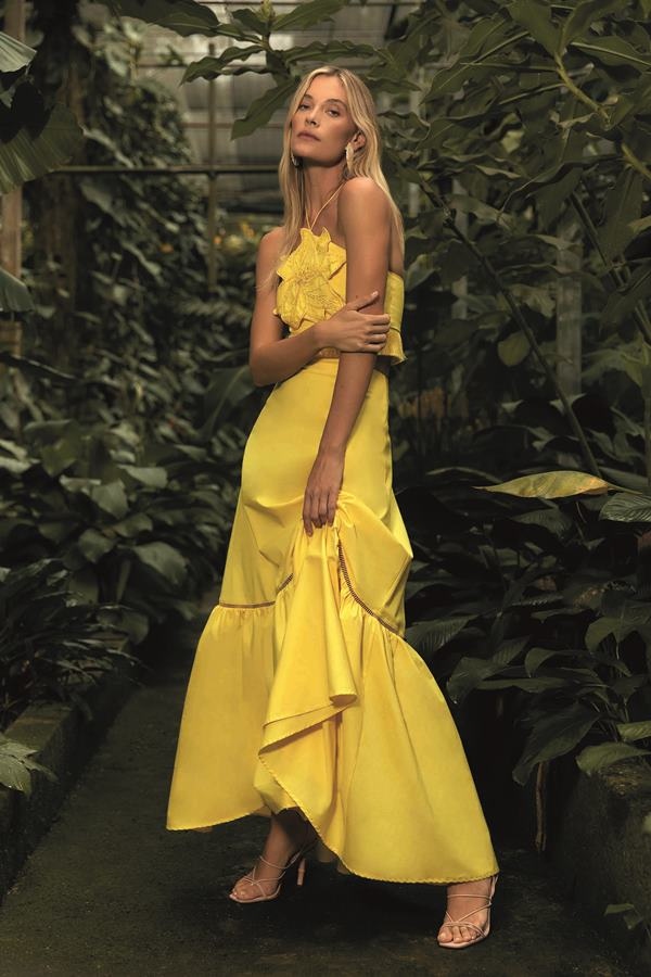 Mulher com vestido amarelo posando para foto. Ela está em frente a uma parece de cerca viva, com plantas - Metrópoles