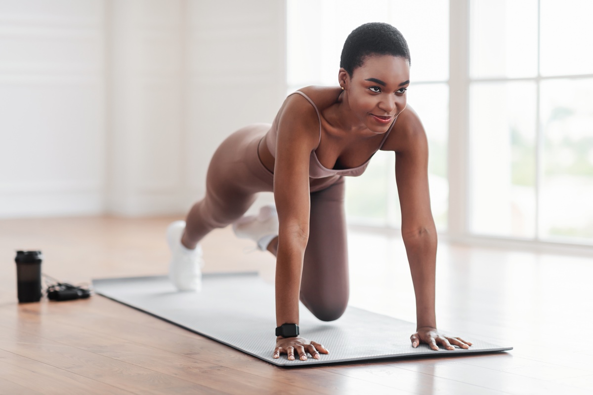 Foto colorida de mulher negra fazendo exercícios com o peso do próprio corpo. Ela está em um tapete - Metrópoles
