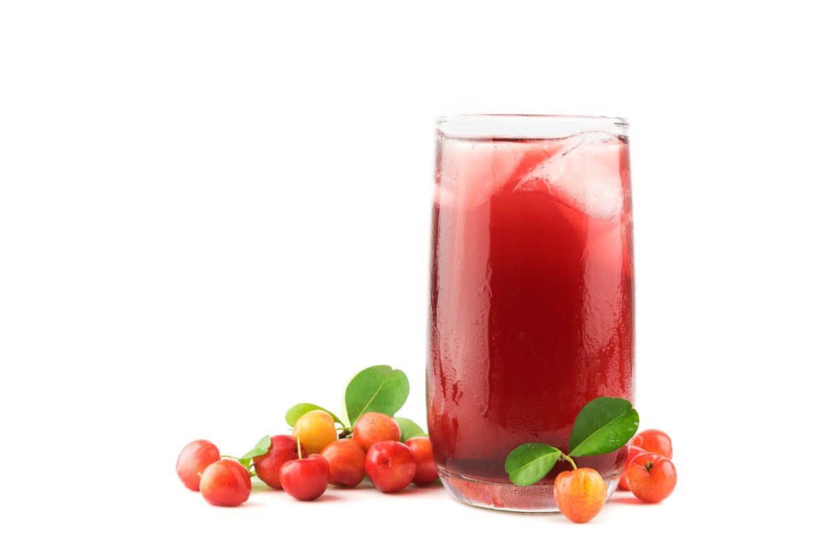 Foto colorida. Em um fundo branco, a imagem mostra um copo transparente com suco de acerola na cor vermelha. Em volta do copo há algumas acerolas - Metrópoles