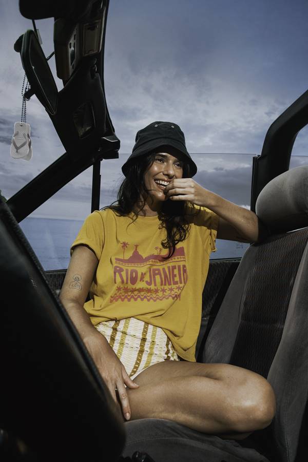 Mulher no carro usando camiseta amarela estampada e chapéu preto - Metrópoels