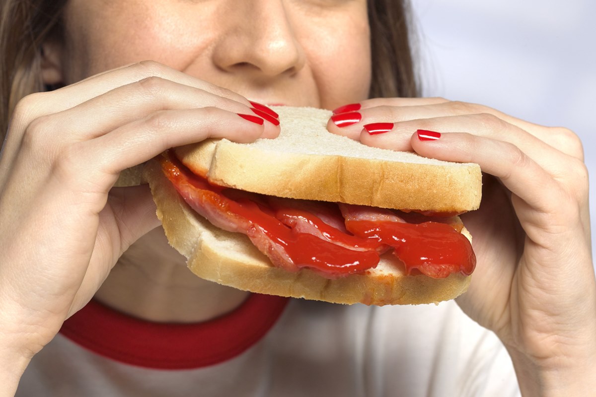 Foto colorida de mulher branca segurando pão com carne processada dentro. As unhas da mulher estão pintadas com esmalte vermelho - Metrópoles