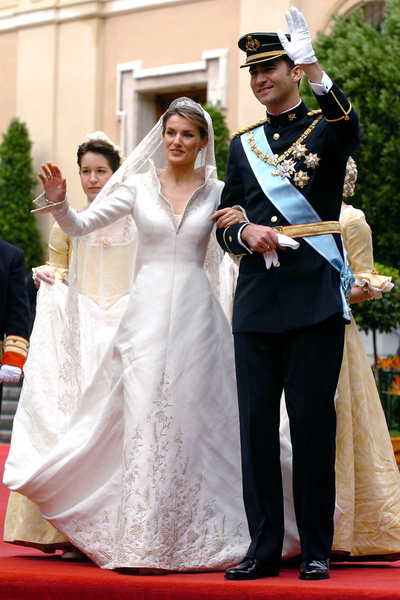 Foto colorida de mulher parda, com roupa de noiva, acenando, enquanto está de mãos dadas com homem vestido com trajes militares - Metrópoles