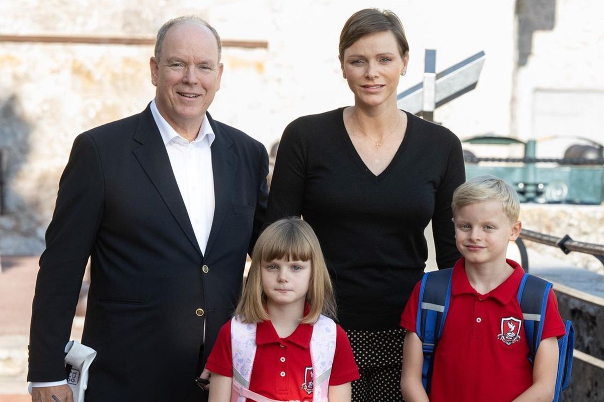Foto colorida de homem e mulher, ambos brancos, com roupas pretas. Na frente deles, tem duas crianças, um menino e uma menina, com uniforme de blusa vermelha - Metrópoles