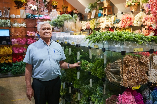 foto colorida de um homem idoso no corredor de uma loja com folhagem de plástico - Merópoles