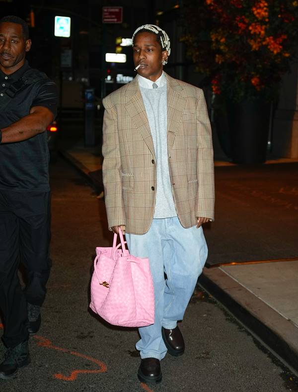 A$ap Rocky usando look com calça jeans e blazer cinza oversized. Ele segura uma bolsa rosa e tem um cigarro na boca - Metrópoles