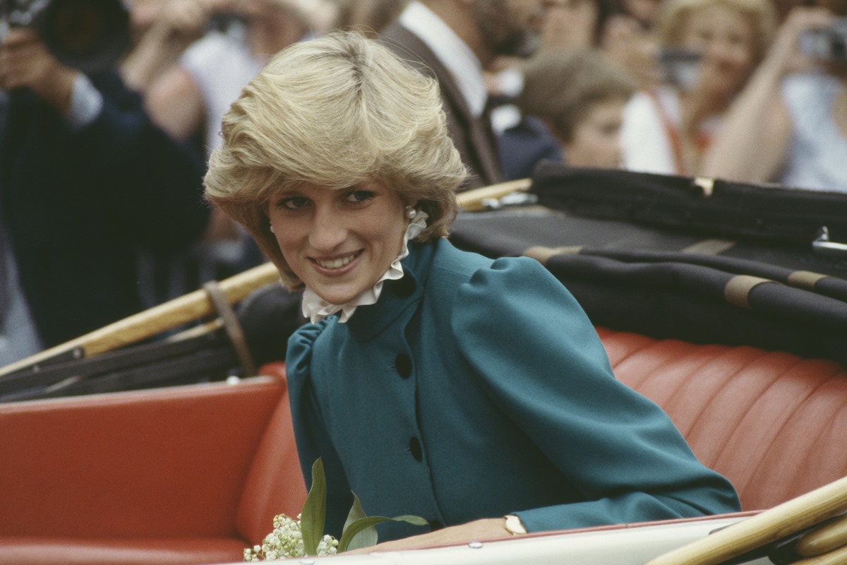Princesa Diana era branca, cabelo loiros curtos e olhos claros. Ela está sentada no carro oficial da realeza com um vestido verde escuro.