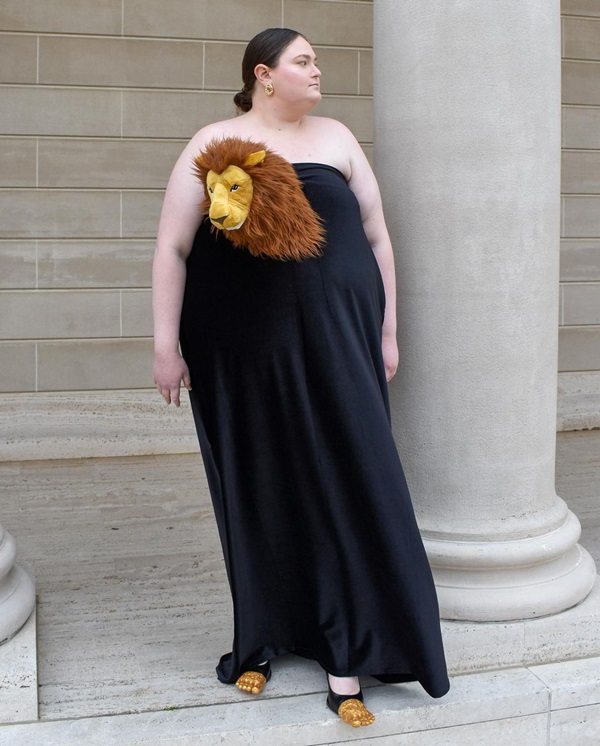 Na imagem com cor, uma mulher branco usando um vestido preto com a cabeça de um leão de pelúclia como fantasia - Metrópoles