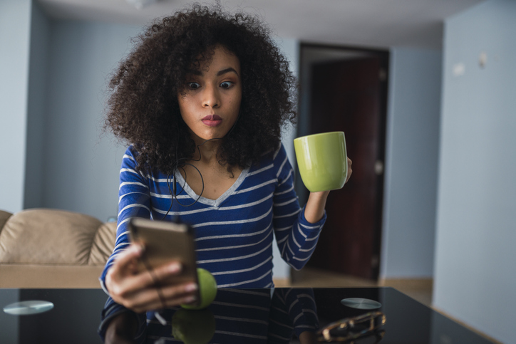 Retrato de jovem com caneca olhando o horário no celular enquanto toma café