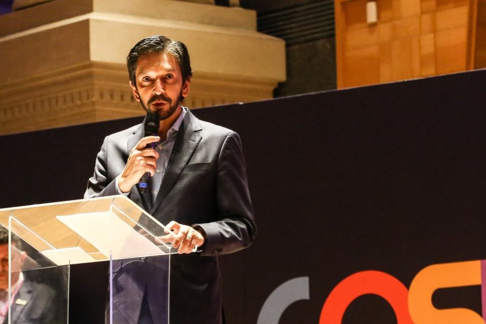 Imagem colorida mostra o prefeito Ricardo Nunes, homem branco, de cabelo e barba pretos, vestindo terno cinza, em um palco. Ele está atrás de um púlpito, falando ao microfone, olhando para o horizonte - Metrópoles