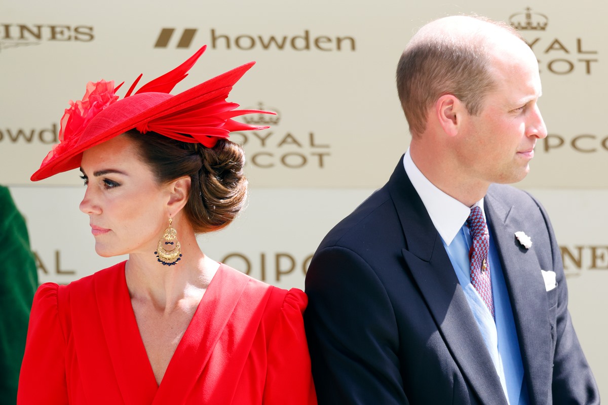 Kate Middleton, mulher branca, de cabelos castanhos, está usando uma roupa e chapéu vermelho. Já o príncipe William está usando um terno preto. Ele está de costas para Kate - Metrópoles