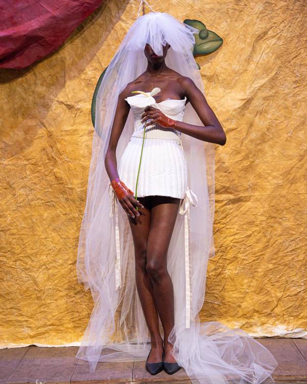 Modelo usando vestido de noiva curto com véu na cabeça e segurando flor - Metrópoles