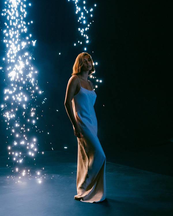 Com luzes de fogos em fundo escuro, Hailey Bieber posa usando vestido branco longo com alcinhas - Metrópoles