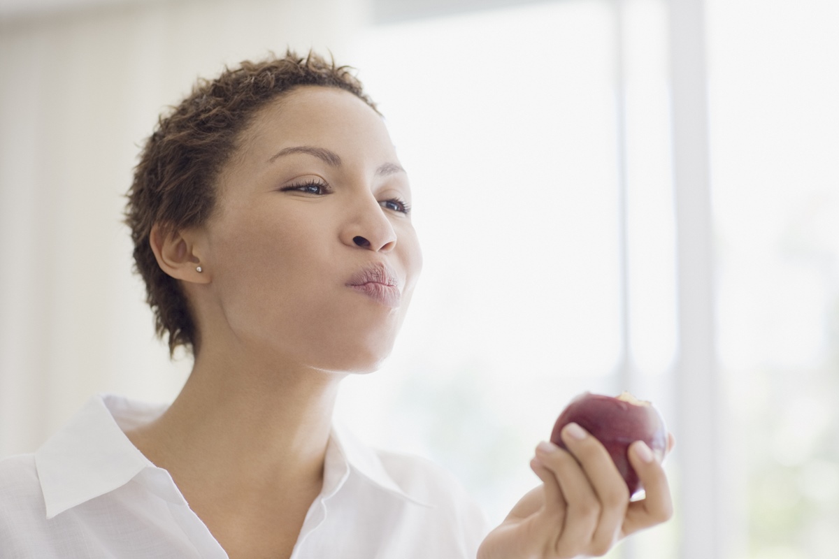 Foto colorida de mulher, com cabelo curto e blusa branca, mastigando uma maçã - Metrópoles