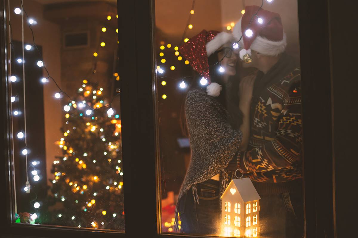 Foto colorida de um casal através da janela na noite de natal com as luzes piscando - Metrópoles