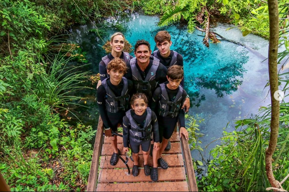 Foto colorida de mulher e homem rodeados por quatro crianças, sendo três meninos e uma menina. Eles estão com roupas de mergulho em uma ponte. Atrás tem um lago - Metrópoles